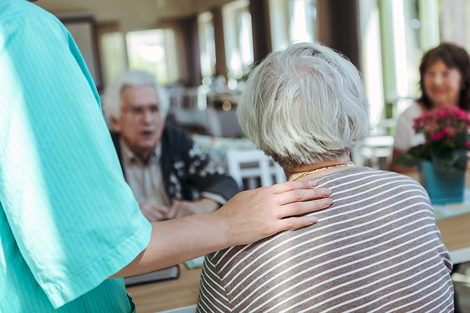 En sjuksköterska lägger handen på en äldre kvinnas axel, i bakgrunden sitter andra personer vid ett bord.