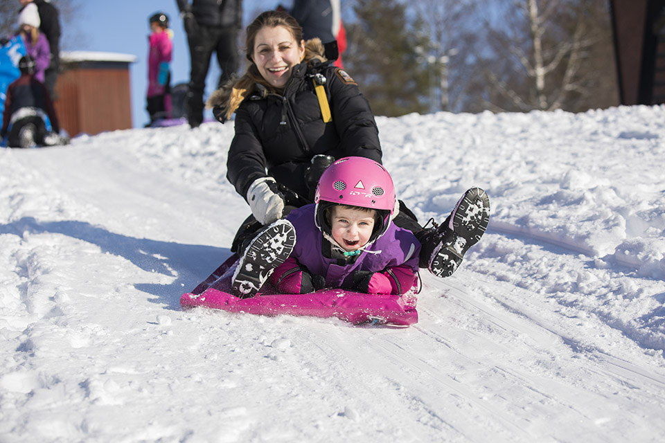 Litet barn med hjälm som åker skrana nedför en pulkabacka, mamma sitter bakom.